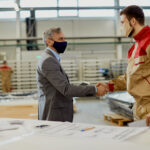 Happy carpenter and businessman shaking hands and greeting in a workshop while wearing protective face masks due to COVID-19 pandemic.