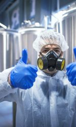 Professional technologist expert in protective white uniform with hairnet, mask and gloves holding both thumbs up. Chemicals